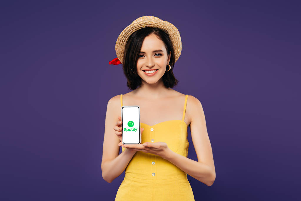 Kijev, Ukrajna-július 3, 2019: mosolygó csinos lány Szalmakalapot gazdaság okostelefon Spotify app izolált lila  - Fotó, kép