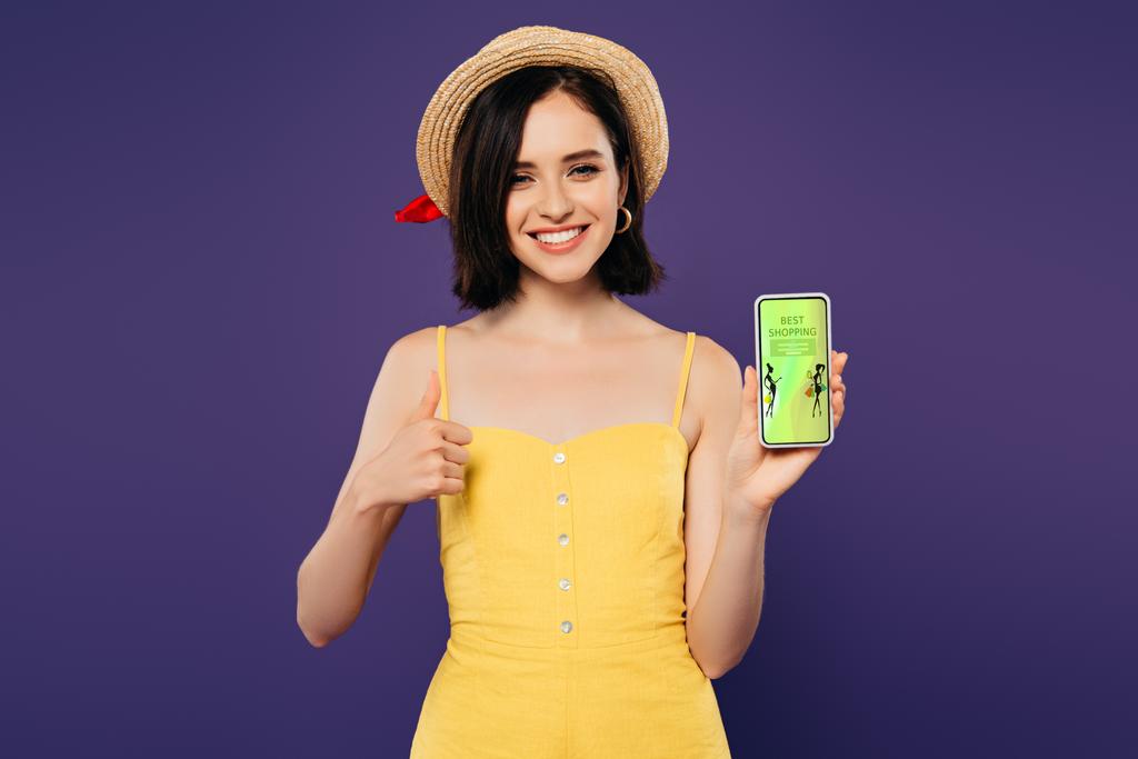 Lächelndes hübsches Mädchen mit Strohhut zeigt Daumen hoch, während es Smartphone mit Mode-Shopping-App in lila hält  - Foto, Bild