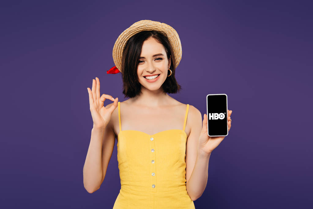 Kiev, Ukrayna - Temmuz 3, 2019: hbo uygulaması ile akıllı telefon tutan hasır şapka güzel kız gülümseyen ve ok işareti mor izole gösteren  - Fotoğraf, Görsel