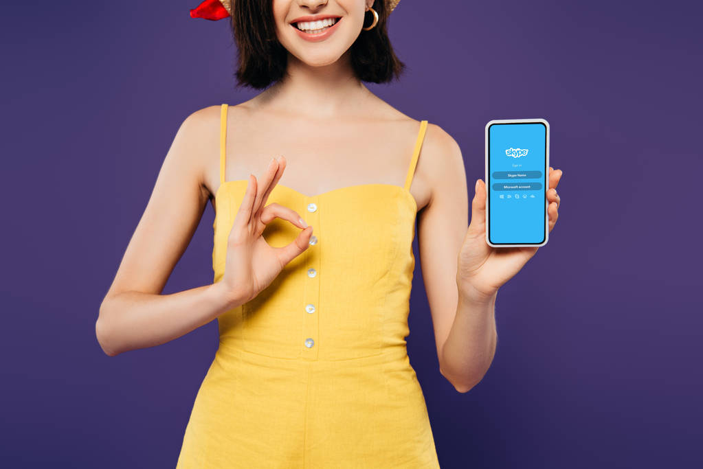 Kijów, Ukraina-3 lipca 2019: przycięte widok uśmiechniętej dziewczyny w gospodarstwie smartphone z logo Skype i pokazując OK znak izolowane na fioletowym  - Zdjęcie, obraz