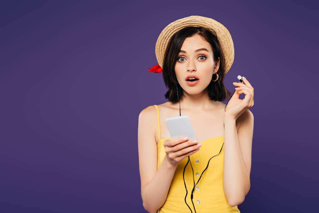 Schockiertes hübsches Mädchen mit Strohhut hört Musik in Kopfhörern auf Smartphone isoliert auf lila - Foto, Bild