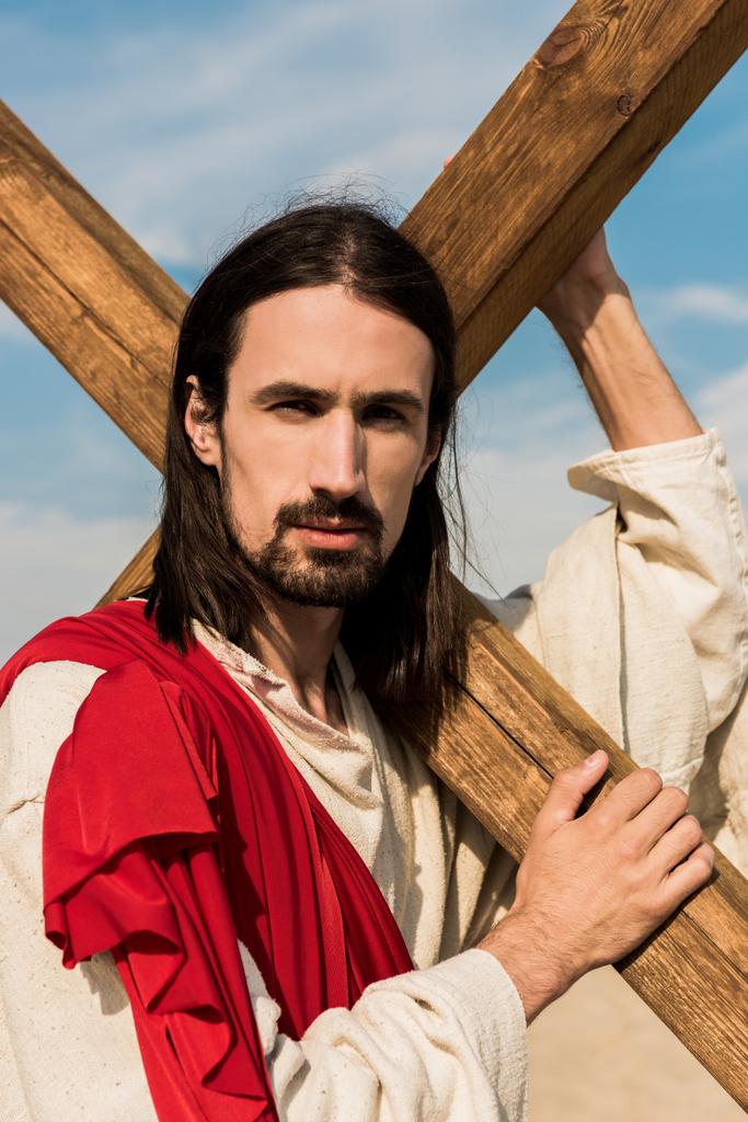 με μούσι Ιησού κρατώντας ξύλινο σταυρό και κοιτάζοντας την κάμερα  - Φωτογραφία, εικόνα