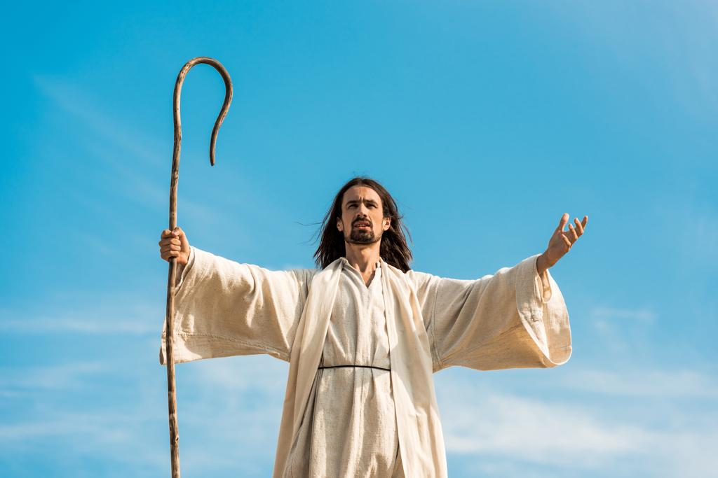 Jesús con las manos extendidas sosteniendo bastón de madera contra el cielo azul
 - Foto, imagen