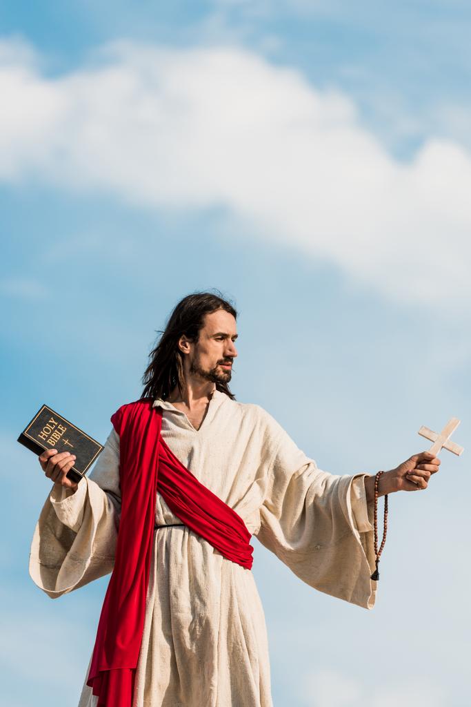 Jesus segurando a Bíblia sagrada e cruz de madeira contra o céu azul com nuvens
  - Foto, Imagem