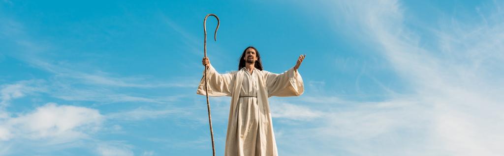 tiro panorâmico de Jesus com as mãos estendidas segurando cana de madeira contra o céu
 - Foto, Imagem