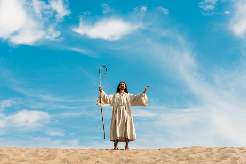 ο Ιησούς με τεντωμένα χέρια κρατώντας ξύλινο μπαστούνι εναντίον του γαλάζιου ουρανού στην έρημο  - Φωτογραφία, εικόνα