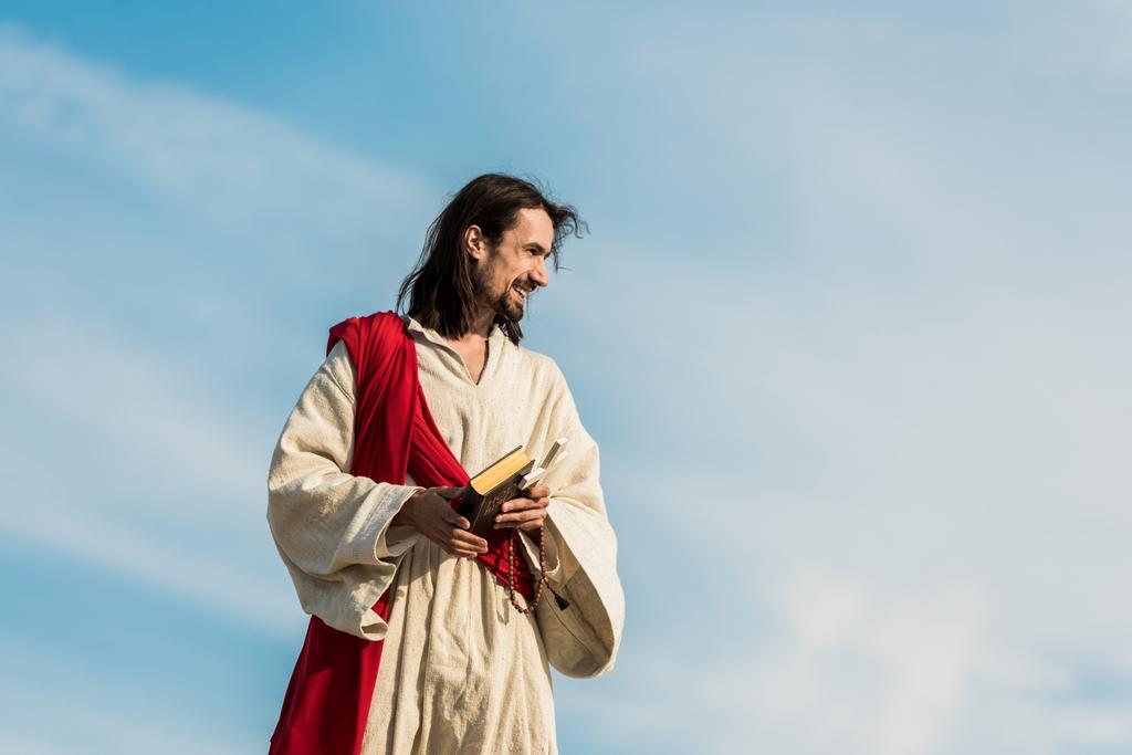 ο Ιησούς κρατώντας σταυρό και ιερή Βίβλο ενάντια στον ουρανό με σύννεφα  - Φωτογραφία, εικόνα