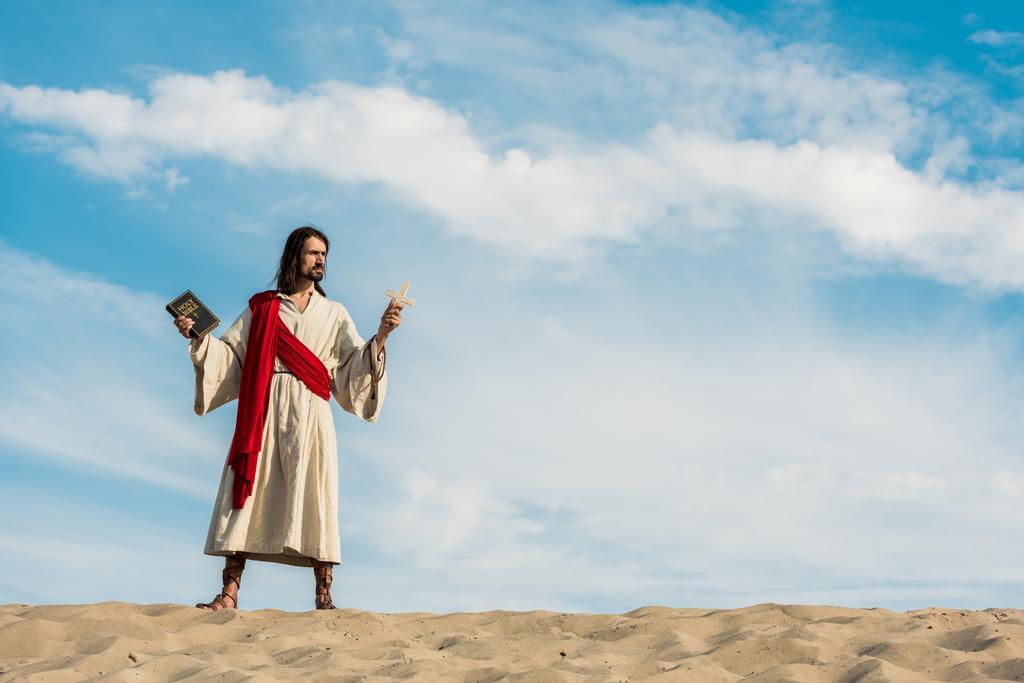 Jezus het houden van de Heilige Bijbel en kruis tegen blauwe hemel met wolken in de woestijn - Foto, afbeelding