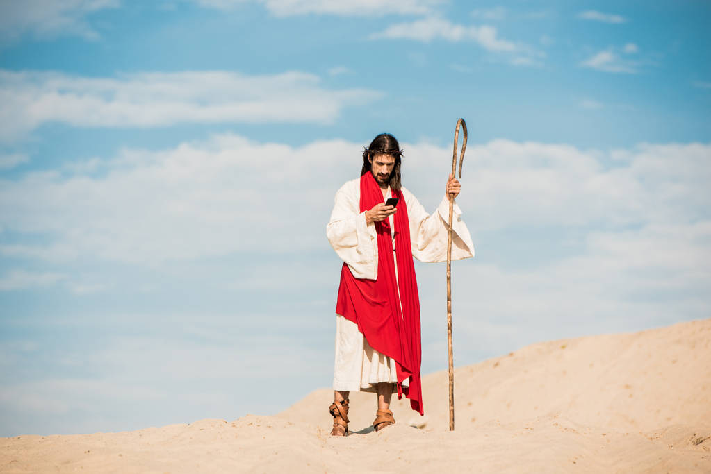 όμορφος άντρας με τον Ιησού μανδύα κρατώντας ξύλινο μπαστούνι και χρησιμοποιώντας smartphone στην έρημο  - Φωτογραφία, εικόνα