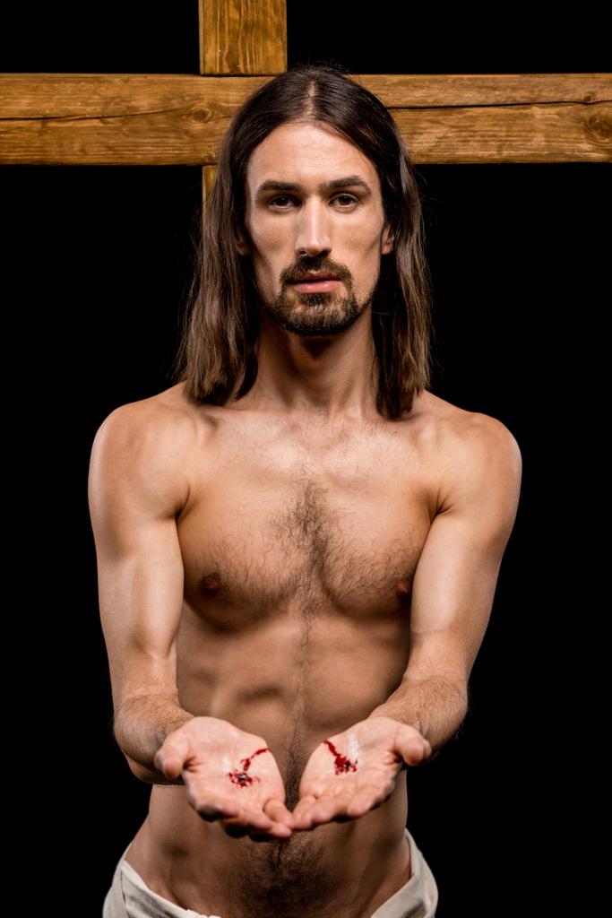 Jezus met bloedige handen staande in de buurt van houten kruis geïsoleerd op zwart  - Foto, afbeelding