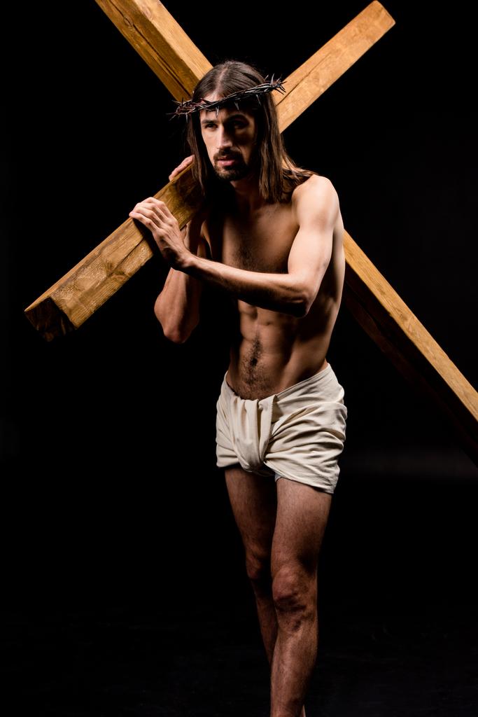 bebaarde shirtless man in krans Holding houten kruis geïsoleerd op zwart  - Foto, afbeelding