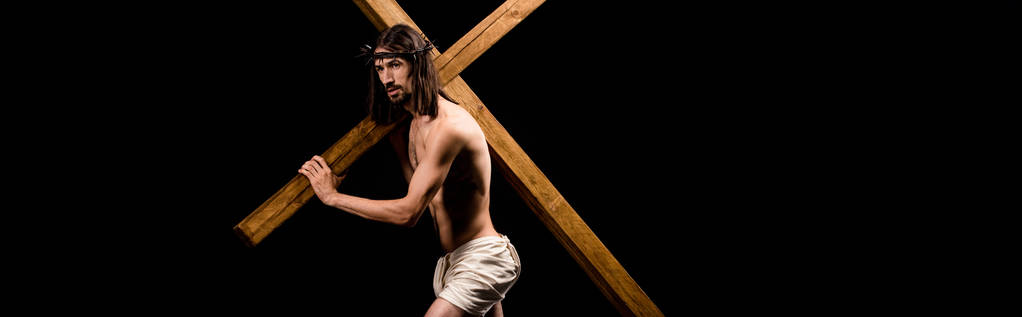 Panoramaaufnahme eines hemdlosen Jesus im Kranz, der ein Holzkreuz hält und isoliert auf schwarz steht  - Foto, Bild