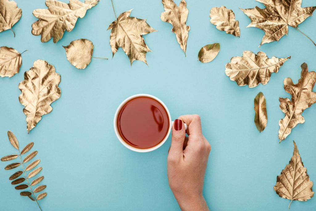 vue recadrée de la femme tenant le thé dans une tasse près du feuillage doré sur fond bleu
 - Photo, image