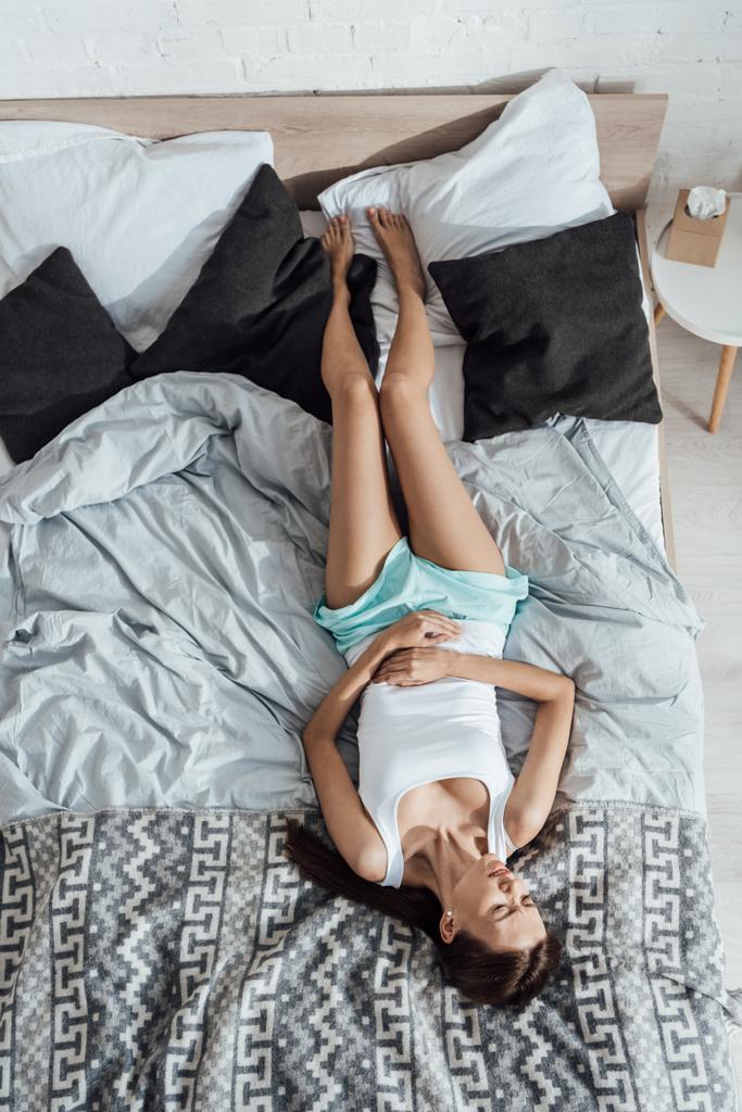 yatakta yatan ve karnına dokunan üzgün genç kadının üst görünümü - Fotoğraf, Görsel