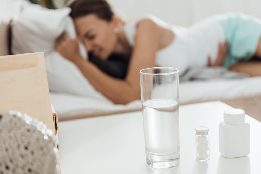 enfoque selectivo de la mujer que sufre en la cama y pastillas con vaso de agua en primer plano
 - Foto, Imagen