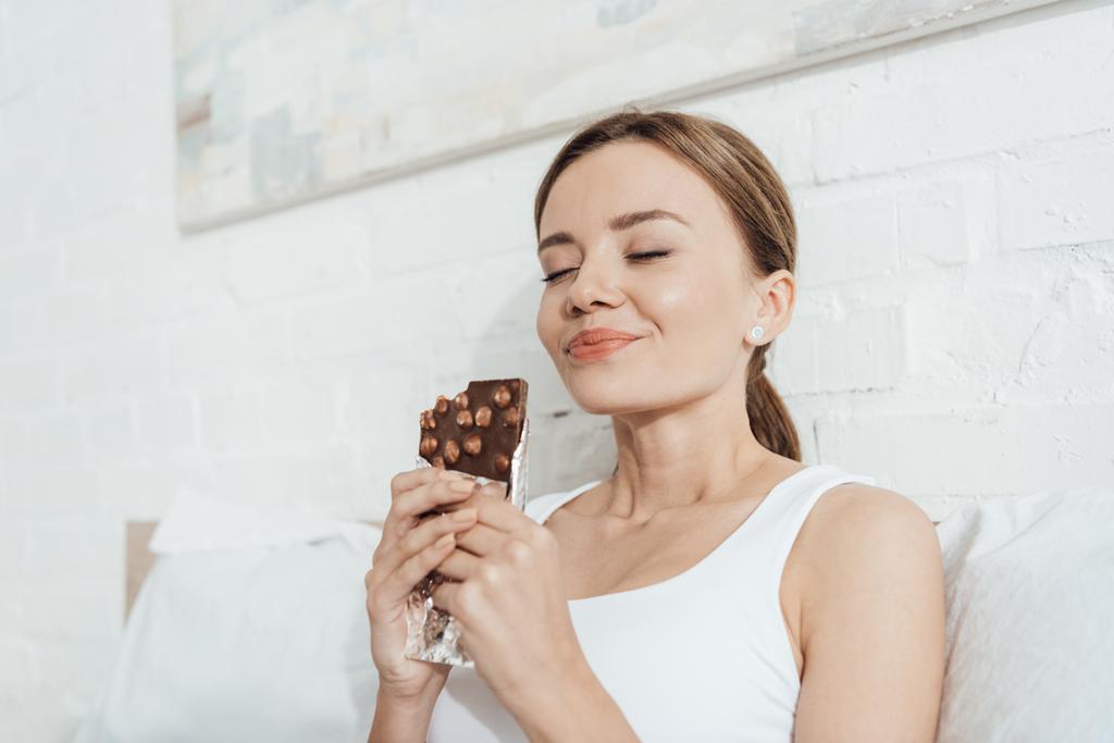 vue à angle bas de la jeune femme souriante mangeant du chocolat aux noix avec les yeux fermés au lit
 - Photo, image