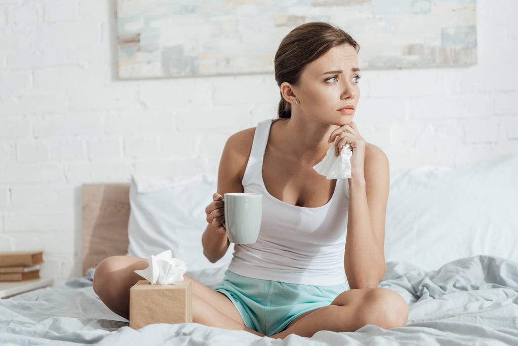 beklemtoonde jonge vrouw zittend op bed en het houden van kopje thee - Foto, afbeelding