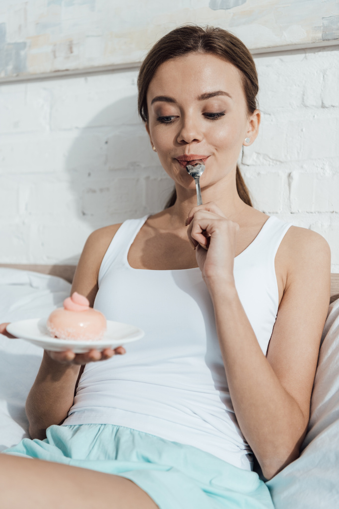 alegre joven mujer acostada en la cama y comiendo cupcake
 - Foto, Imagen
