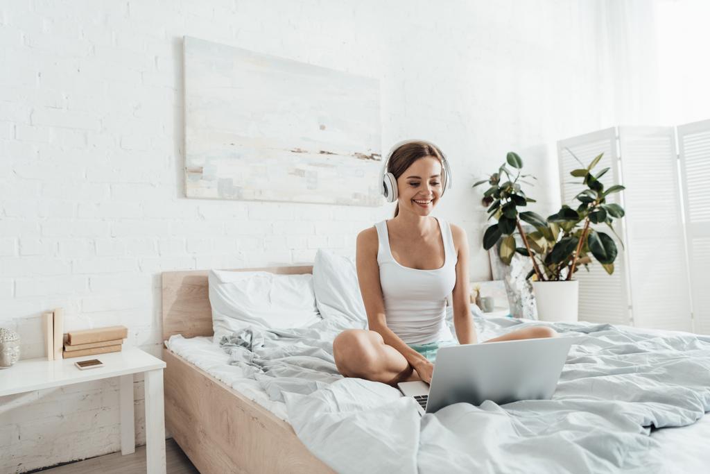 smiling young woman listening music in headphones and using laptop on bed - Photo, Image