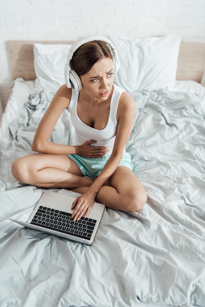 mujer joven en auriculares tocando el vientre y el uso de ordenador portátil en la cama
 - Foto, imagen
