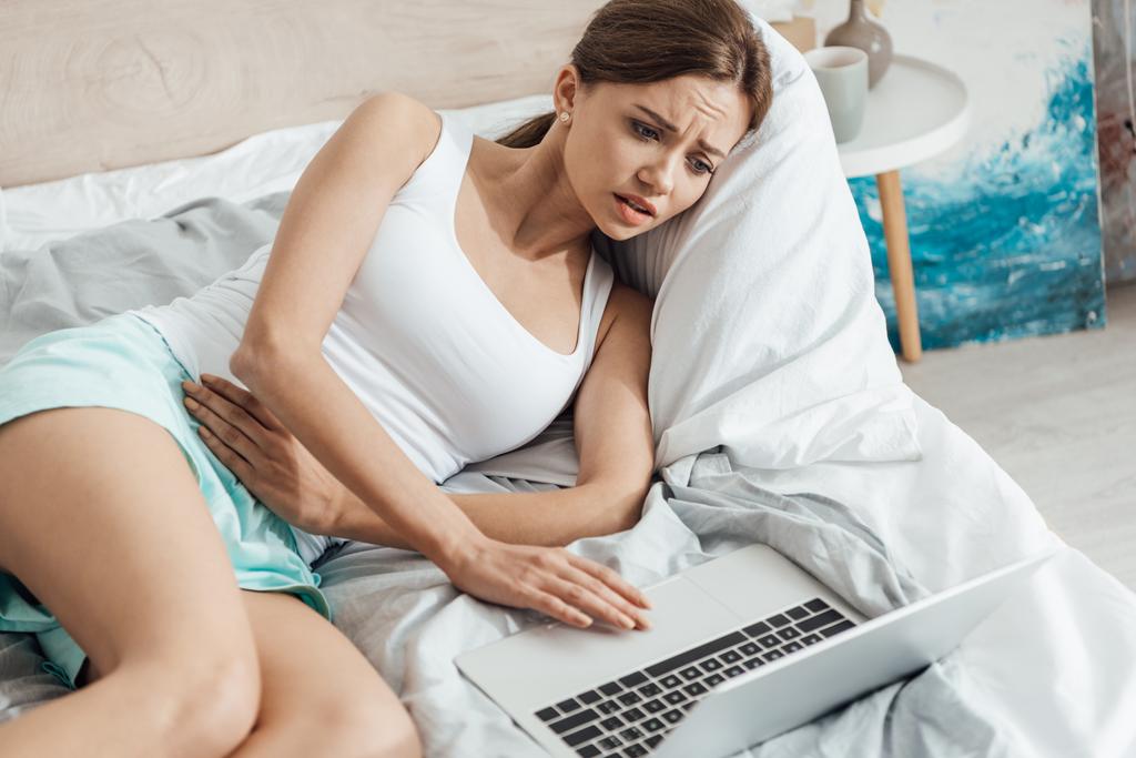 Junge Frau liegt mit Laptop im Bett und berührt Bauch - Foto, Bild