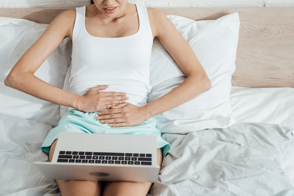 visão parcial da mulher estressada tocando barriga ao usar laptop na cama
 - Foto, Imagem