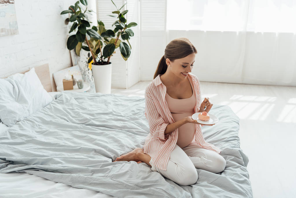 усміхнена вагітна жінка сидить на ліжку і тримає тарілку з кексом
 - Фото, зображення