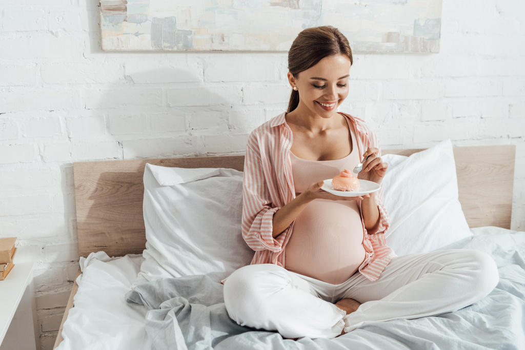 uśmiechnięta kobieta w ciąży siedzi na łóżku i trzyma spodka z Cupcake - Zdjęcie, obraz