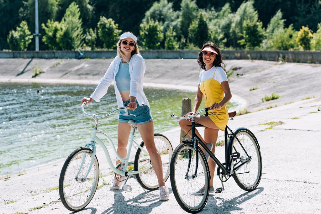 glückliche blonde und brünette Mädchen auf Fahrrädern in Flussnähe im Sommer - Foto, Bild