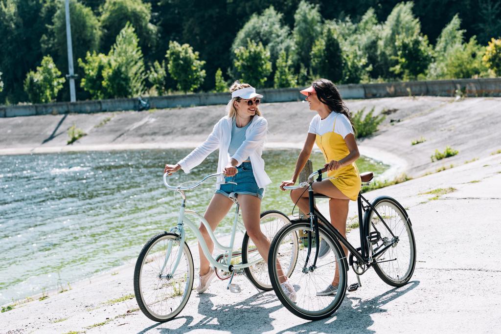 счастливые блондинки и брюнетки с велосипедами, смотрящие друг на друга у реки летом
 - Фото, изображение
