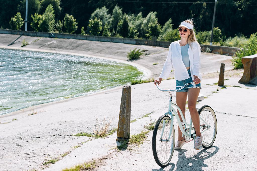 夏に川の近くで自転車に乗ってブロンドの美しい女の子 - 写真・画像