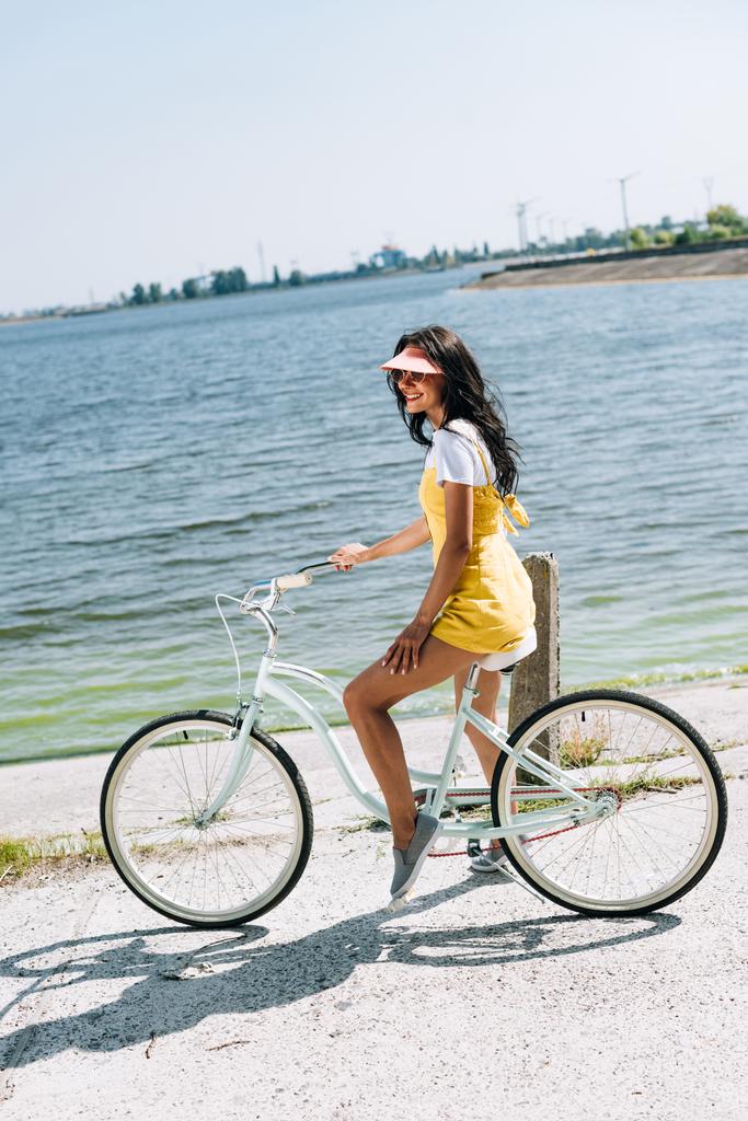 Вид сбоку счастливой брюнетки, катающейся на велосипеде у реки летом
 - Фото, изображение