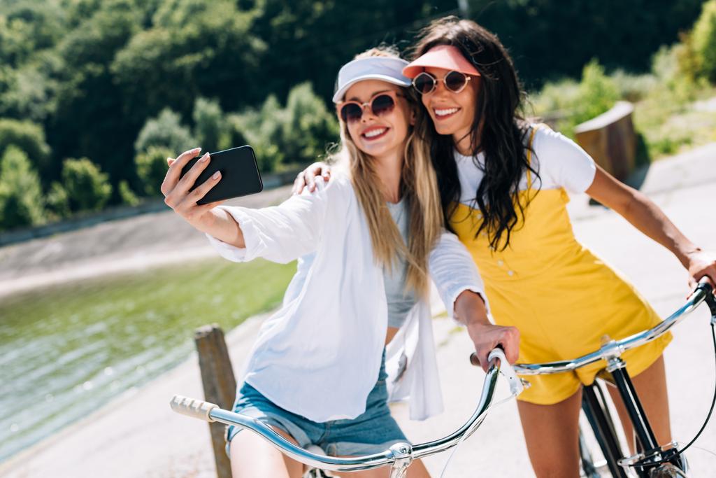 selective focus of blonde and brunette girls riding bikes and taking selfie in summer - Photo, Image