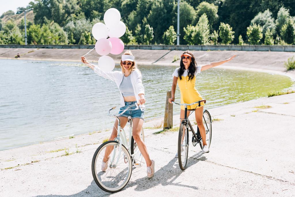Gelukkig blonde en brunette meisjes rijden fietsen met ballonnen in de buurt van de rivier in de zomer - Foto, afbeelding