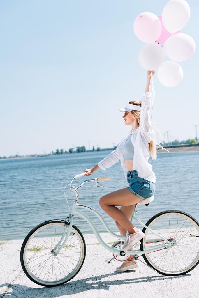 pohled na usměvavou blondýnku jezdijící na kole s balónky poblíž řeky v létě - Fotografie, Obrázek