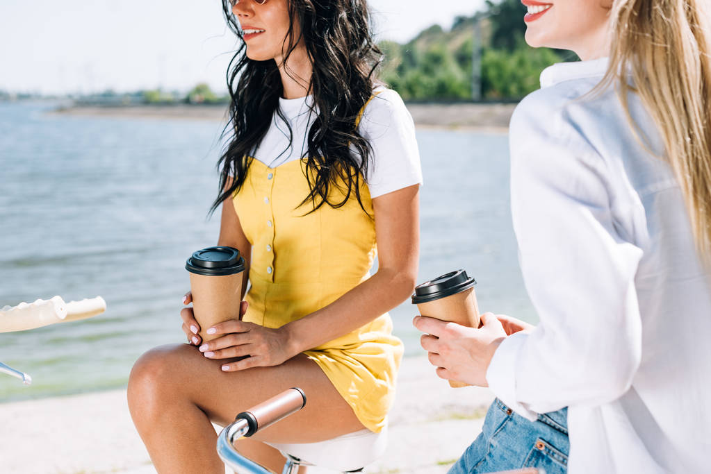 cropped view of girls on bikes with coffee to go near river in summer - Photo, Image