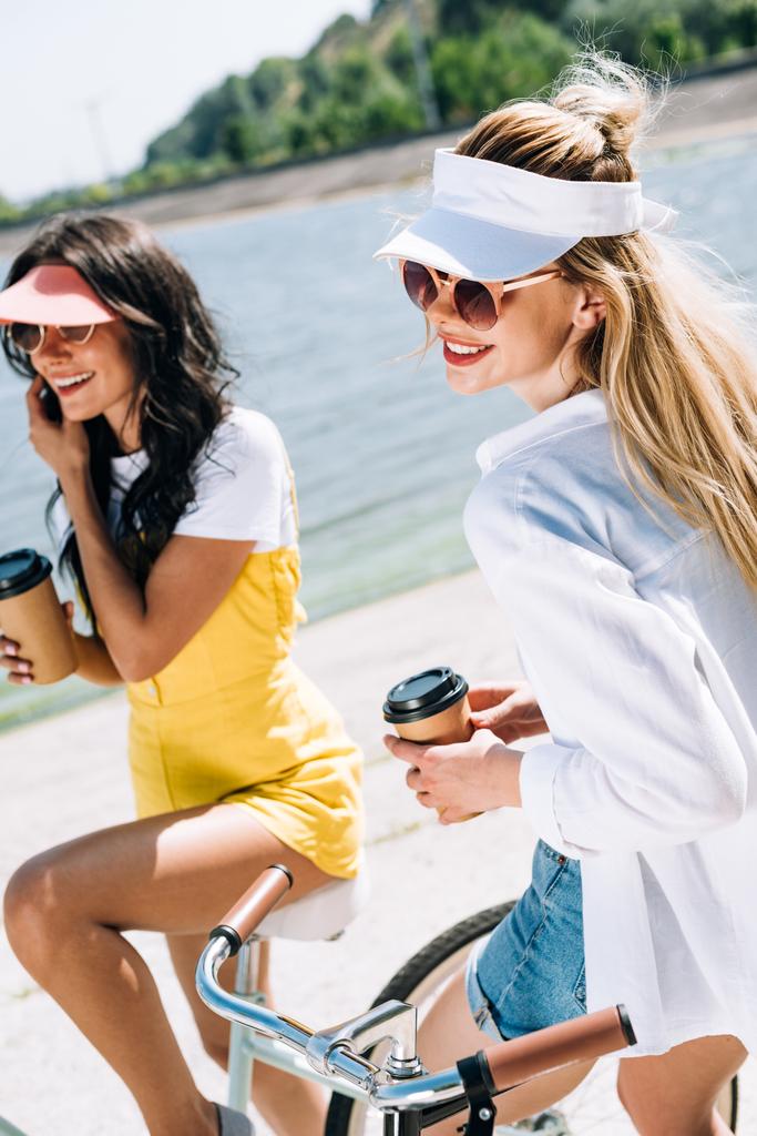 happy blonde and brunette friends on bikes with coffee to go near river in summer - Photo, Image