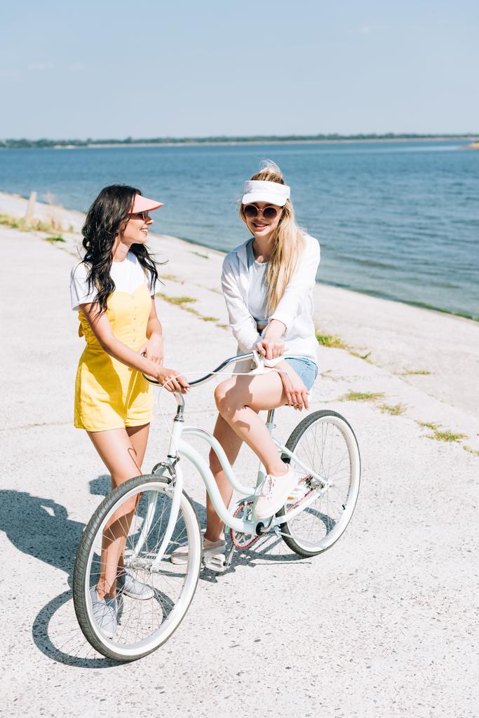 usmívající se blondýnka a brunetka s cyklistou poblíž řeky v létě - Fotografie, Obrázek