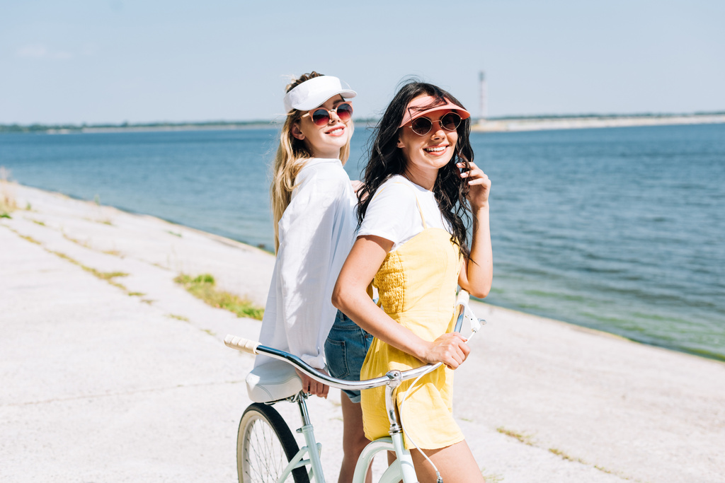 uśmiechnięta Blondynka i Brunette Dziewczyny z rowerem w pobliżu rzeki w lecie - Zdjęcie, obraz