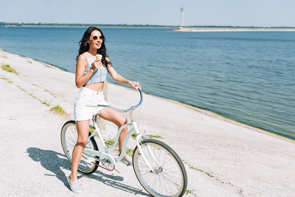 smiling brunette girl with ice cream riding bike near river in summer - Photo, Image
