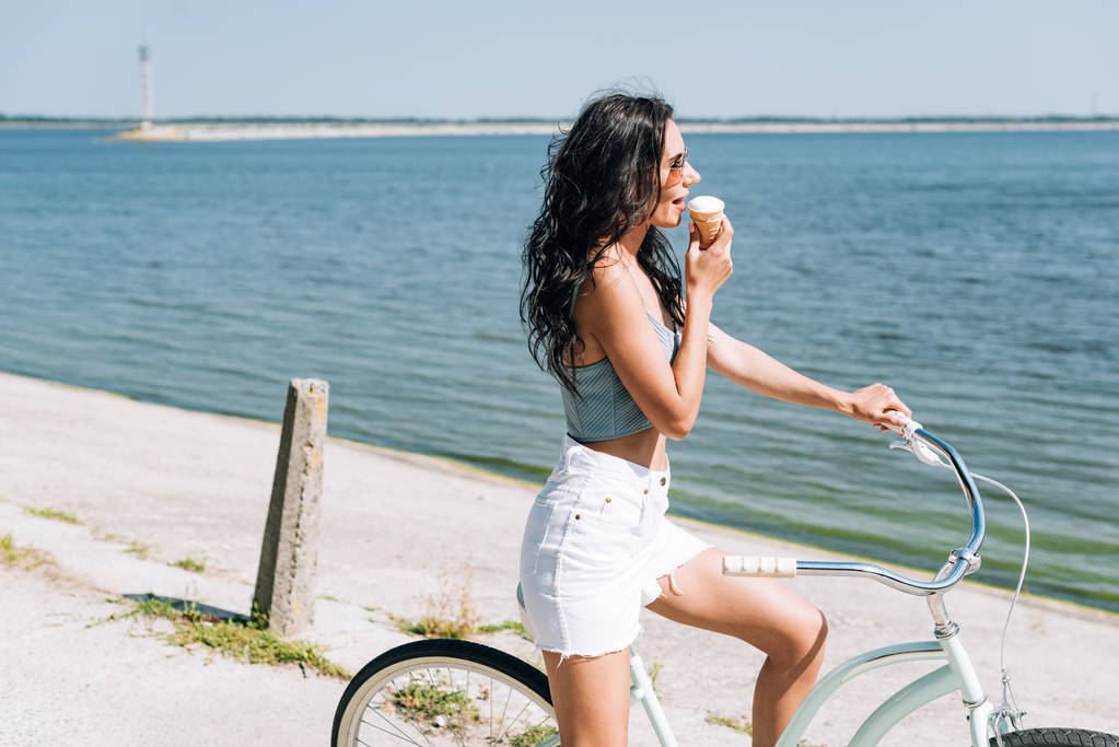 Seitenansicht des brünetten Mädchens, das Eis isst und im Sommer in Flussnähe Fahrrad fährt - Foto, Bild