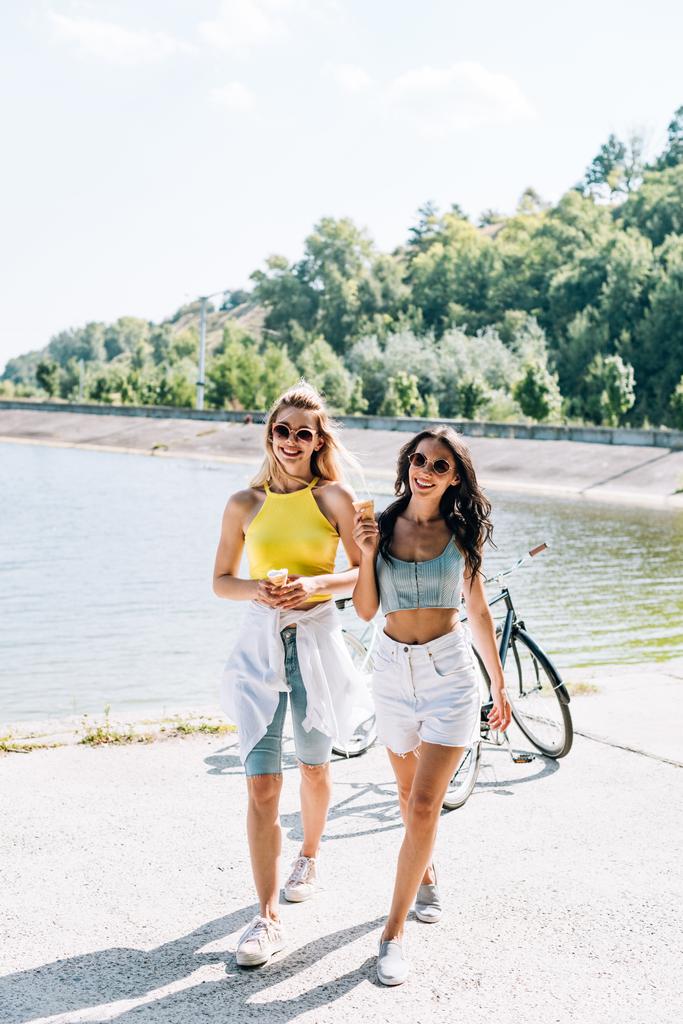 chicas rubias y morenas felices cerca de bicicletas con helado en el río en verano
 - Foto, Imagen