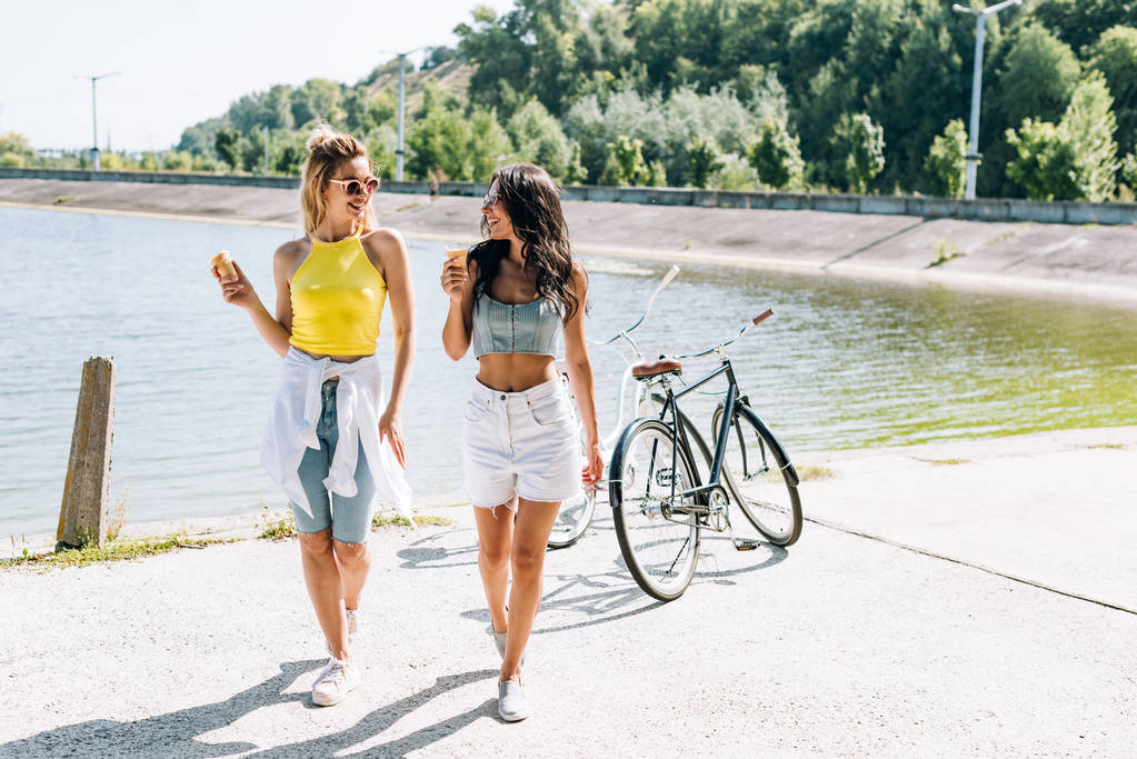 夏に川でアイスクリームと自転車の近くに笑顔ブロンドとブルネットの女の子 - 写真・画像