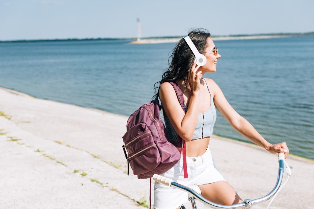 sırt çantası bisiklet ve yaz aylarında nehir yakınında kulaklık müzik dinleme ile mutlu kız - Fotoğraf, Görsel