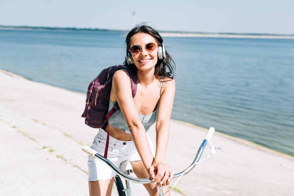 Uśmiechnięta dziewczyna z plecaka rowerowych i słuchania muzyki w słuchawkach w pobliżu rzeki w lecie - Zdjęcie, obraz