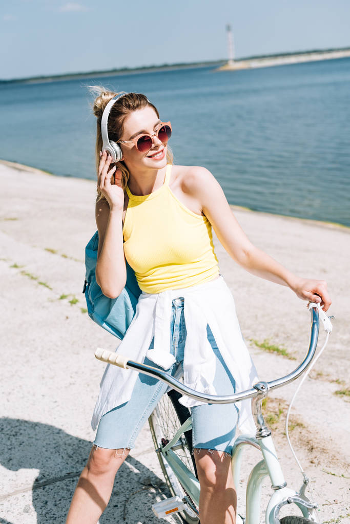 Blond dziewczyna z plecaka rowerowych i słuchania muzyki w słuchawkach w pobliżu rzeki w lecie - Zdjęcie, obraz