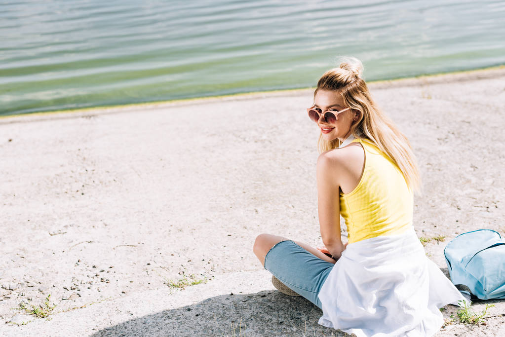 yaz aylarında nehir yakınında oturan sırt çantası ile sarışın mutlu kız - Fotoğraf, Görsel