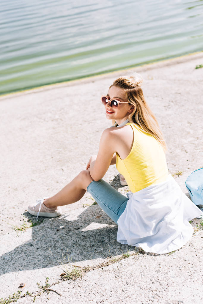 bionda ragazza felice in occhiali da sole con zaino seduto vicino al fiume in estate
 - Foto, immagini