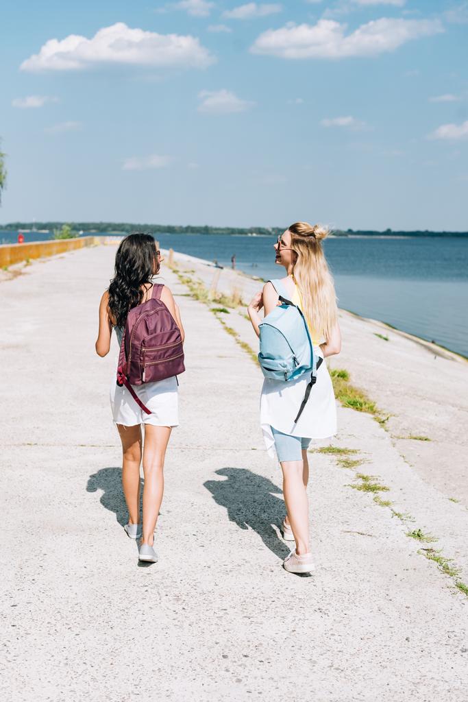 back view of girls walking with backpacks near river in summer - Photo, Image