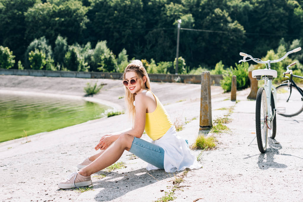 yaz aylarında nehir yakınında oturan bisiklet ile gülümseyen sarışın kız - Fotoğraf, Görsel